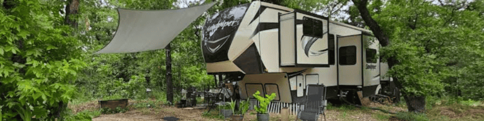 Elgin TX RV Park Elgin RV Park Colinas Texas wooded campsite with parked RV, chairs, potted plants, shade canopy, gravel ground, and tall trees.